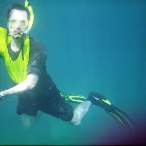 A snorkeler wearing a yellow vest and fins poses underwater while holding a sea scooter and mesh collection bag in clear blue-green water.
