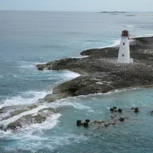 An aerial view of a small white lighthouse on a rocky point surrounded by turquoise and teal ocean waves breaking against the shore.
