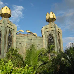 Two ornate stone towers with golden lotus-shaped domes rise above tropical palm trees against a partly cloudy blue sky.