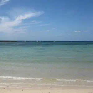 A pristine white sand beach with crystal-clear turquoise water gently lapping at the shore under a blue sky with wispy clouds.