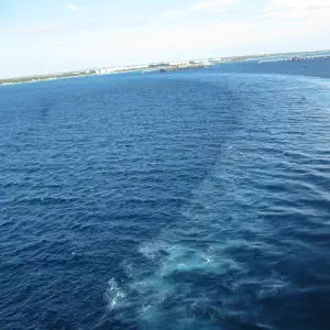 View from a cruise ship railing looking out at deep blue ocean water with a white wake trail curving away from a port town visible on the horizon.