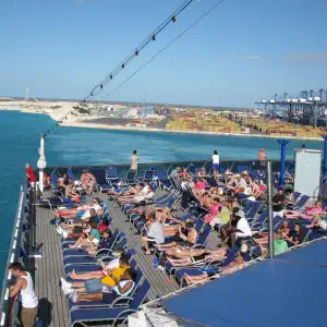 Cruise ship sun deck crowded with passengers lounging on blue deck chairs as the ship passes a port with cranes and shipping containers visible in the background.