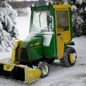A green and yellow John Deere 318 garden tractor with an enclosed cab and front-mounted snowblower attachment parked in a snowy driveway surrounded by snow-covered evergreen trees.