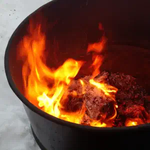 Bright orange flames burn inside a dark metal fire pit sitting on snow-covered ground.