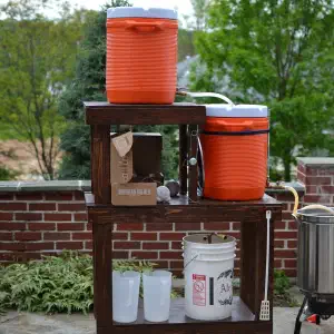A wooden outdoor beverage cart with two orange coolers on top, a burner and pot on the side, and storage shelves below, positioned on a stone patio with brick pillars and green trees in the background.