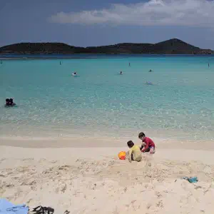 Children and swimmers enjoying a pristine turquoise beach with white sand, dark volcanic islands visible across the water under a blue sky with white clouds.