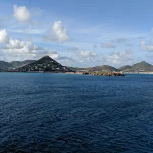 A view from the water of a mountainous Caribbean island with scattered white buildings along the hillsides under partly cloudy skies.