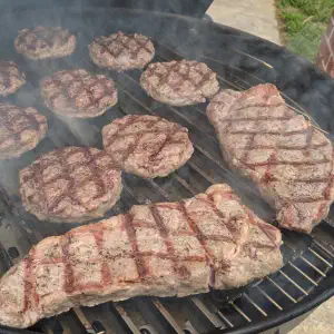 A black grill with hamburger patties and steaks cooking over charcoal, with smoke rising from the surface.