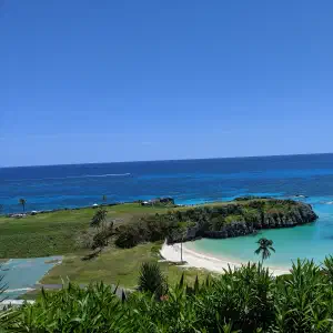 An elevated view of a turquoise cove with a small sandy beach, rocky cliffs, palm trees, and green lawns meeting the deep blue Atlantic Ocean.