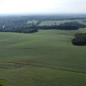 An aerial drone photo of expansive green farmland with rolling hills, tree lines, and distant mountains under a hazy sky.