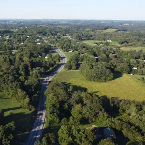 An aerial drone photograph of a two-lane road winding through rolling green farmland with scattered houses and trees stretching to the horizon.