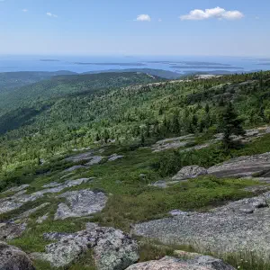 A panoramic view from a rocky granite summit overlooking lush green forested hills stretching to the ocean under a blue sky with scattered clouds.