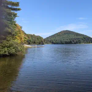A calm lake reflecting a tree-covered hill with early fall foliage in yellows and greens, a dock visible on the far shore under a clear blue sky.