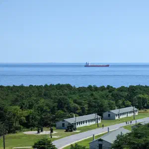 An elevated view of military barracks-style buildings on green lawns with a cargo ship passing through the Delaware Bay under a clear blue sky.