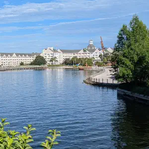 Disney's Yacht Club Resort viewed across a calm lake with lush green trees framing the white New England-style buildings under a blue sky.