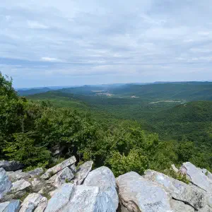 A view from a rocky outcrop over a vast green forested valley with rolling hills under a partly cloudy sky.