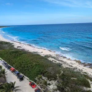 An aerial view of a curved white sand beach and turquoise ocean with a line of colorful Jeeps parked along a coastal road bordered by green vegetation.