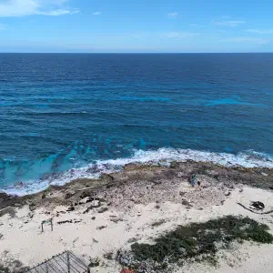 An elevated view of a rocky shoreline meeting turquoise Caribbean waters with a sandy beach and a few visitors below.
