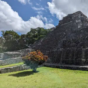 A large stepped Mayan stone pyramid surrounded by green grass and tropical trees under a dramatic cloudy sky.