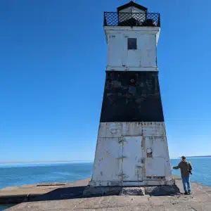 A person standing next to a weathered black and white lighthouse at the end of a concrete pier with calm blue water and clear sky.