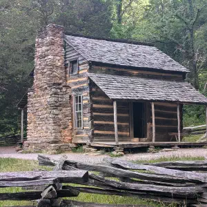 A rustic log cabin with a stone chimney surrounded by mature forest trees, with weathered wooden fence rails in the foreground.