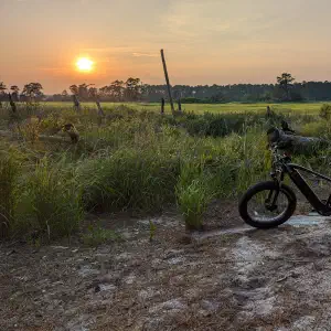 Fat tire electric bike parked on a sandy trail beside a lush green salt marsh with the sun setting through hazy skies