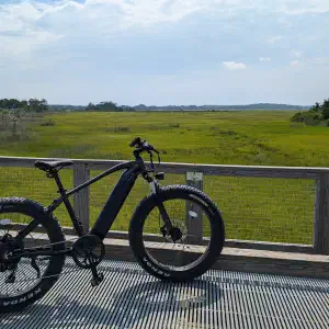 Fat tire electric bike on a wooden boardwalk observation deck overlooking a vast green salt marsh under a blue sky with scattered clouds