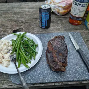 A plated meal on a rustic wooden table with potato salad and green beans on one side, a grilled steak on a cutting board on the other, accompanied by a beer can, whiskey bottle, and snack bags.