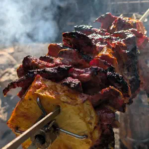 Spit-roasted meat with charred edges on vertical skewers over a fire, with smoke rising in the background.