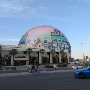 The Las Vegas Sphere displaying a Dead and Company concert advertisement at dusk with palm trees and a blue sports car in the foreground.