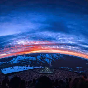 Fisheye view from inside the Las Vegas Sphere showing a concert stage surrounded by crowds with a dramatic sunset over snow-capped mountains displayed on the wraparound screen