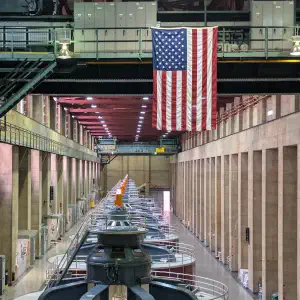 Interior of a hydroelectric power plant facility with large turbine generators lined up in a row, concrete walls on either side, industrial equipment overhead, and an American flag hanging from the ceiling.