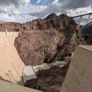 The Hoover Dam's massive concrete curved structure with rugged canyon walls and rocky terrain beneath a partly cloudy sky.