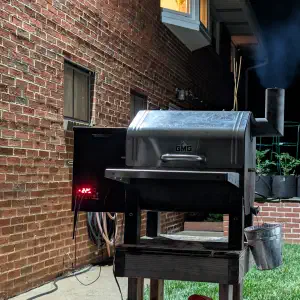 Green Mountain Grills pellet smoker with smoke rising from the chimney at night on a brick patio with a garden of potted plants in the background