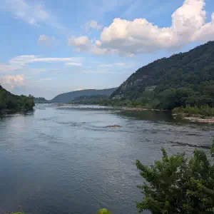 A wide river flowing between two steep forested hillsides under a partly cloudy sky with cumulus clouds.