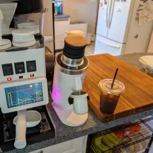 Gaggiuino Gaggia Classic espresso machine with a live shot profiling graph on its touchscreen next to a conical burr grinder and an iced coffee on a walnut cutting board