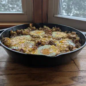 A cast iron skillet filled with diced potatoes, ground meat, and eggs cooking on a wooden floor in front of windows.