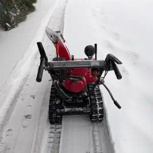 A red and black snow blower photographed from above clearing a snowy driveway lined with evergreen trees.