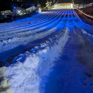A nighttime overhead view of an illuminated snow tubing slope with parallel white snow lanes lit by blue lighting, flanked by golden railings and lodge buildings.