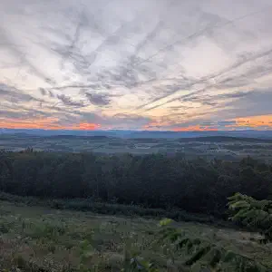 A sweeping landscape view at sunset showing forested ridges in the foreground and a vast patchwork valley below, with dramatic orange and purple clouds stretching across the sky.