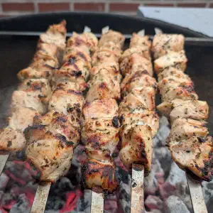 Five seasoned meat skewers grilling over hot coals and flames in a metal barbecue pit.