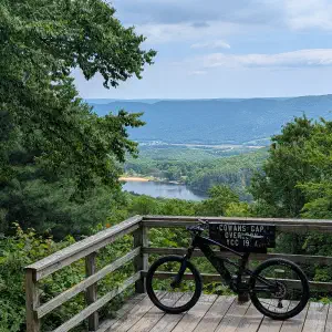 A black bicycle leans against a wooden railing on an overlook platform, with a scenic vista of forested mountains, a lake, and valley landscape in the distance.